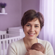 Close-up portrait of a smiling young mother, mid-20s, holding a baby, in a soft-focus nursery with a dusty lilac and white ba